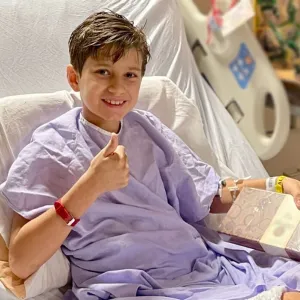 Boy sits in hospital bed, giving a thumbs up