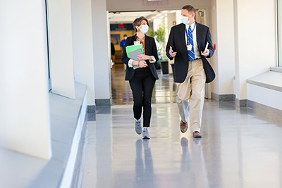 Gerry Grant walks down a hallway on the Duke Medical Center Campus, talking to Kate McDaniel