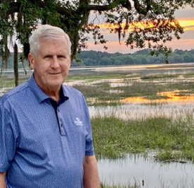 A man smiles outdoors in front of trees and a lake at sunset.