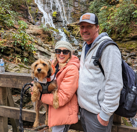 A woman stands in front of a waterfall with a man and their dog. They are smiling.