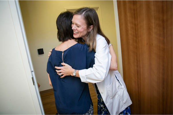 A doctor in a white coat smiles and hugs her patient.