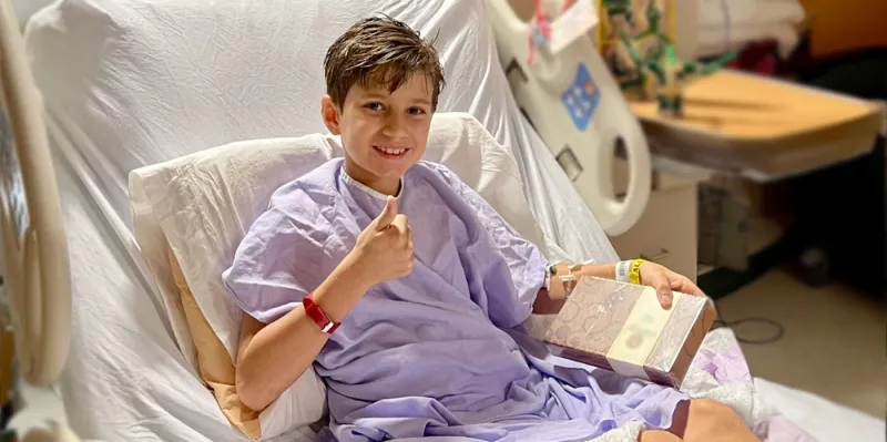 Boy sits in hospital bed, giving a thumbs up