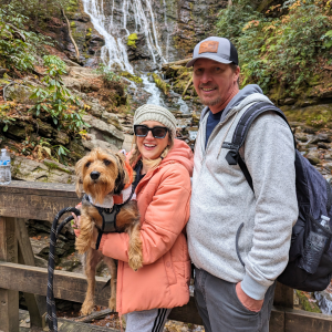 Heather Gensler holds her dog, Ryder, while her husband, Bobby, stands next to her. They are on a bridge in front of a waterfall.